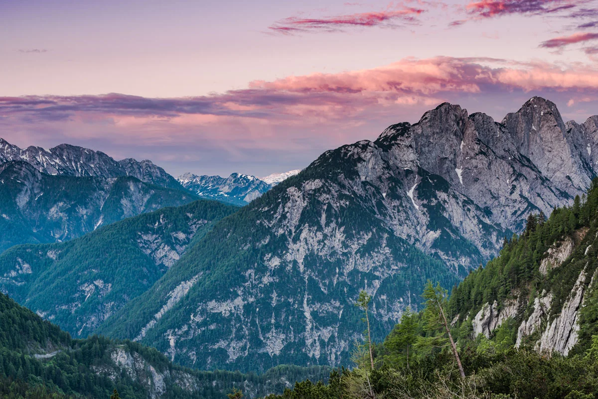 Sončni zahod nad vrhovi Julijskih Alp s pogledom na gozdnate strmine in rožnato obarvano nebo