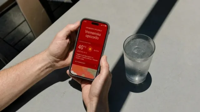 Pogled od zgoraj na roke, ki držijo pametni telefon z rdečim vremenskim opozorilom, zraven pa stoji kozarec ledene vode na mizi.